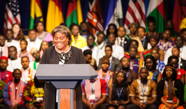 Eine Frau, die an einem Rednerpult mit Mikrofonen spricht und einer aufmerksamen Zuhörerschaft gegenübersteht, mit sichtbaren Flaggen im Hintergrund.