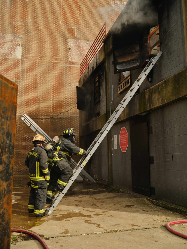 Feuerwehrleute in Helmen klettern an einer Leiter zu einem rauchgefüllten Gebäude hoch, mit einem Metallobjekt links und Rohren auf dem Boden darunter.