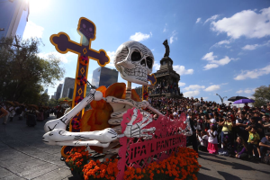Ein Totentag-Umzug in Mexiko-Stadt zeigt eine zentrale Skelettstatue mit Blumen und Kreuz, umgeben von Menschen mit Schirmen, die gehen oder sitzen, sowie Gebäuden, Bäumen und einem bewölkten Himmel im Hintergrund.