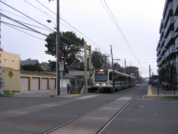 Ein Nahverkehrszug fährt durch eine Straße mit hohen Gebäuden, Strommasten und einigen Fußgängern, mit Bäumen und einem klaren blauen Himmel im Hintergrund.