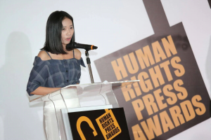 Eine Frau in einem schwarzen Kleid steht an einem Rednerpult mit einem Mikrofon und hält eine Rede bei den Human Rights Press Awards, mit einem ernsten und konzentrierten Gesichtsausdruck.