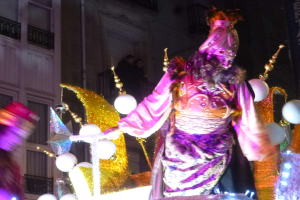 Ein Mann in einem Kostüm steht auf einem dekorierten, beleuchteten Festwagen in einer Stadtparade, mit einem mehrstöckigen Gebäude mit Fenstern und Geländern im Hintergrund.