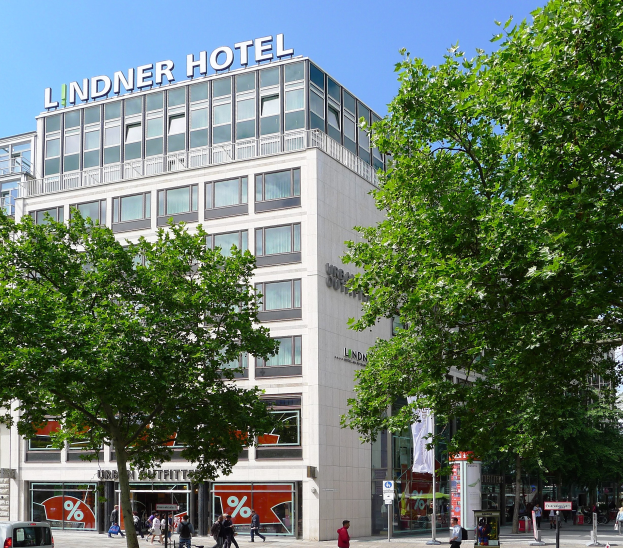 Großes modernes Hotelgebäude mit Glasfenstern, das Wort "Lindner" oben drauf, umgeben von Bäumen, Fahrzeugen und Fußgängern unter einem sichtbaren Himmel.