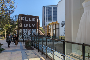 Menschen gehen auf einem Gehweg vor einem Gebäude mit einer Tafel, auf der "Elle Bully" steht, vorbei. Rechts sind Bäume, Pflanzen und eine Schaufensterpuppe zu sehen. Im Hintergrund gibt es weitere Gebäude und einen klaren blauen Himmel.