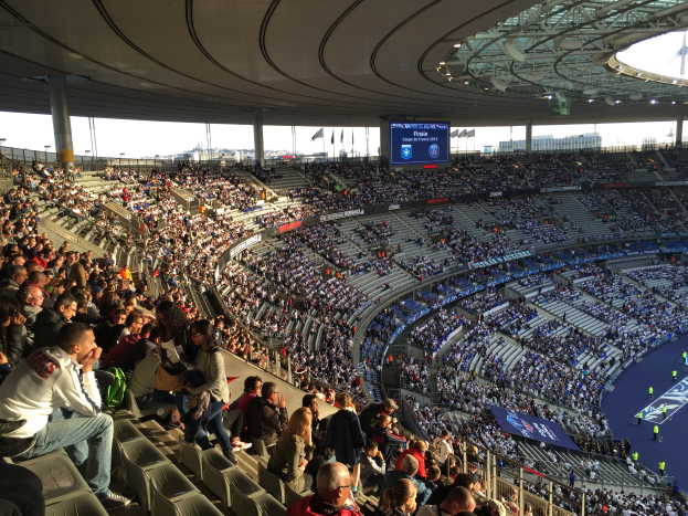 Eine große Menschenmenge sitzt im Allianz Stadion in München, Deutschland, bei einem Fußballspiel, mit einer Bühne auf der rechten Seite und Fahnen, Stangen und einem Bildschirm im Hintergrund.