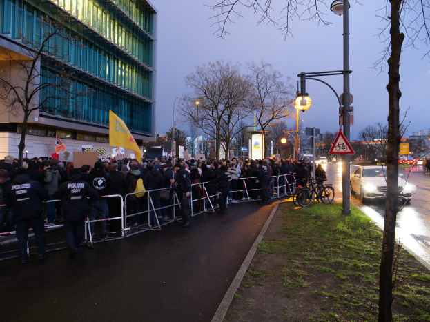 Eine große Gruppe von Menschen mit Schildern steht vor einem Gebäude mit Barrikaden, Fahrrädern, Laternen, Schildern, Bäumen und Gras, mit dem Himmel im Hintergrund, was auf eine Protestaktion in Berlin hinweist.