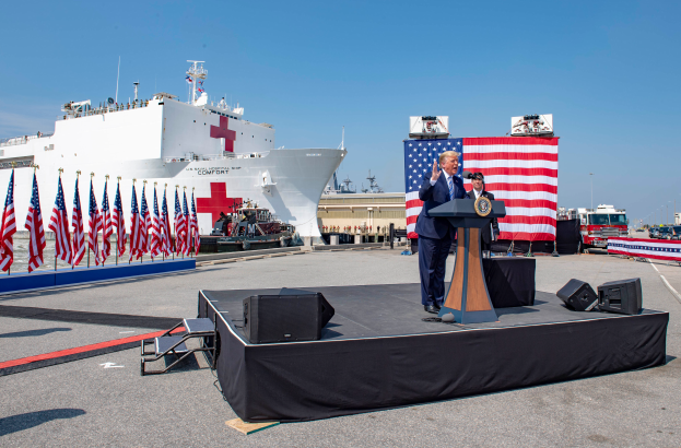 Präsident Donald Trump spricht an einem Rednerpult vor einem großen Schiff, umgeben von Flaggen, Fahrzeugen und anderen Gegenständen, trägt ein Sakko, eine Krawatte und ein Hemd und hält ein Mikrofon in der Hand, mit dem Himmel im Hintergrund.