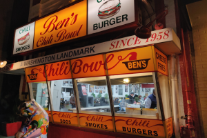 Ein Gebäude mit Glaswänden und beleuchteter Beschilderung, das ein paar Menschen im Inneren und eine Wand mit Rohren auf der rechten Seite zeigt.