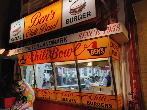 Ein Gebäude mit Glaswänden und beleuchteter Beschilderung, das ein paar Menschen im Inneren und eine Wand mit Rohren auf der rechten Seite zeigt.