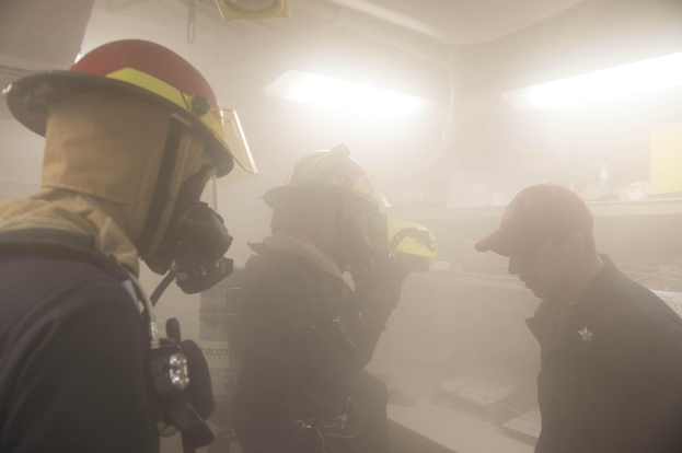 Feuerwehrleute mit Helmen und Rauch vor dem Mund, einer hält eine Kamera, während einer Trainingsrunde mit Hintergrundbeleuchtung und -gegenständen.
