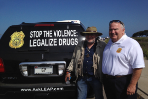 Zwei Männer stehen neben einem schwarzen Van mit der Aufschrift "Stop the Violence Legalize Drugs"; einer hält eine Flasche, mit Menschen, Bäumen und einem klaren blauen Himmel im Hintergrund.