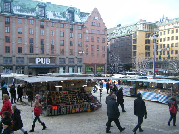 Ein lebendiger Weihnachtsmarkt in Stockholm, Schweden, mit Menschen, die spazieren gehen, Ständen voller Gegenstände, Gebäuden, Laternenmasten, Bäumen und einem klaren blauen Himmel im Hintergrund.