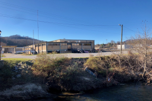 Ein Fluss fließt durch eine grüne Wiese neben einem Gebäude, mit Bäumen, Pflanzen, Gras, Steinen, Straßenlaternen, Strommasten, Stromkabeln und Hügeln unter einem klaren blauen Himmel an der Stelle einer geplanten Wasseraufbereitungsanlage.