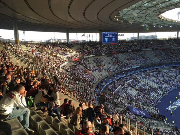 Große Zuschauermenge im Allianz Arena Stadion bei einem Fußballspiel, mit einer Bühne, Fahnen, Stangen und einem Bildschirm im Hintergrund.
