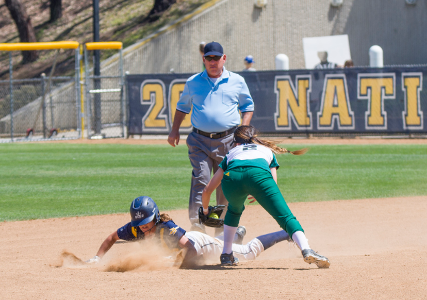 Ein Baseballfeld mit drei Personen auf rotem Boden in der Mitte, Bäumen, Einzäunung, einer Wand, einem Banner mit Text und anderen Gegenständen im Hintergrund.