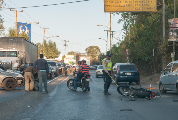 Eine Gruppe von Menschen steht um ein verungl√ľcktes Motorrad am Stra√üenrand mit mehreren Fahrzeugen, darunter ein Lastwagen, und einer Hintergrund von B√§umen, Masten, Lichtern und Schildern unter dem Himmel.