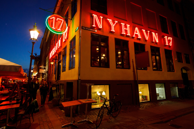 Ein Gebäude mit rotem Text an seiner Fassade, begleitet von einem Fahrrad, Tischen und Stühlen, mit einer Straßenansicht daneben.