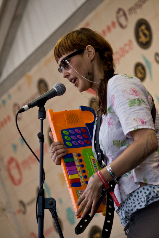 Eine Frau in einem weißen Hemd und Sonnenbrille singt in ein Mikrofon, während sie ein Piano spielt, mit einem Plakat an der Wand dahinter.