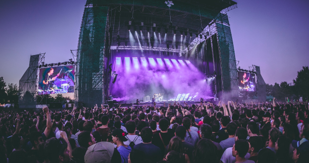 Eine große Menschenmenge auf einem Musikfestival mit einer Bühne in der Mitte, auf der Musiker Instrumente spielen, beleuchtet von Scheinwerfern und umgeben von Bildschirmen, unter einem klaren blauen Himmel mit einem Gebäude und Bäumen im Hintergrund.