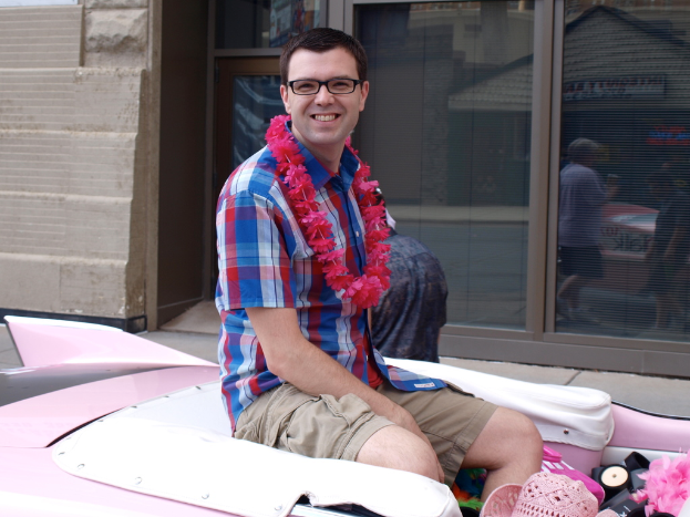 Ein Mann mit Brille sitzt und lächelt in der Mitte, mit einem Fahrzeug unten, einem Gebäude, Glastüren und Gehweg im Hintergrund, das Menschen, Gebäude und Schilder reflektiert.
