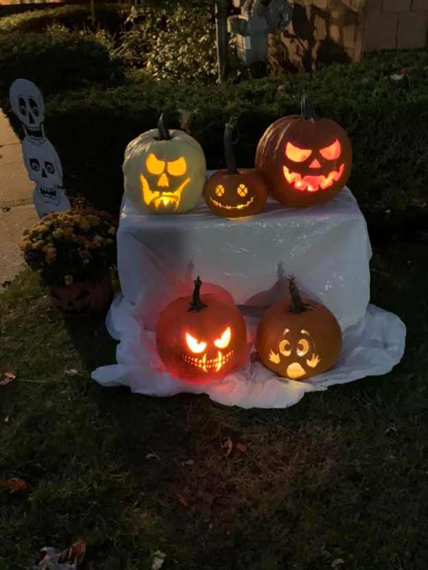 Eine Gruppe von geschnitzten Kürbissen auf einem weißen tuchbedeckten Tisch, umgeben von Gras und trockenen Blättern, mit Pflanzen, Bäumen und einer Wand im Hintergrund.