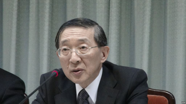 Ein Mann im Anzug und Krawatte sitzt an einem Tisch mit einem Mikrofon, trägt eine Brille und befindet sich in einem formellen Setting mit einem Vorhang im Hintergrund, während die Hand einer anderen Person links im Bild zu sehen ist.