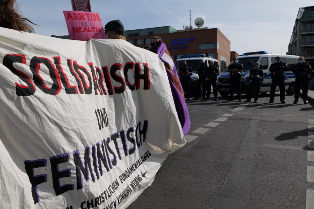 Gruppe von Menschen, die eine Straße entlanggehen und eine 'Solidarität und Feminismus'-Schlagzeile halten, mit geparkten Fahrzeugen, Gebäuden, einer Schüsselantenne und einem klaren blauen Himmel im Hintergrund.