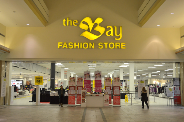 Außenansicht eines Bay Fashion Stores in einem Einkaufszentrum mit Einkäufern, Wandtexttafeln, Deckenleuchten, Säulen und Hintergrundauslagen.