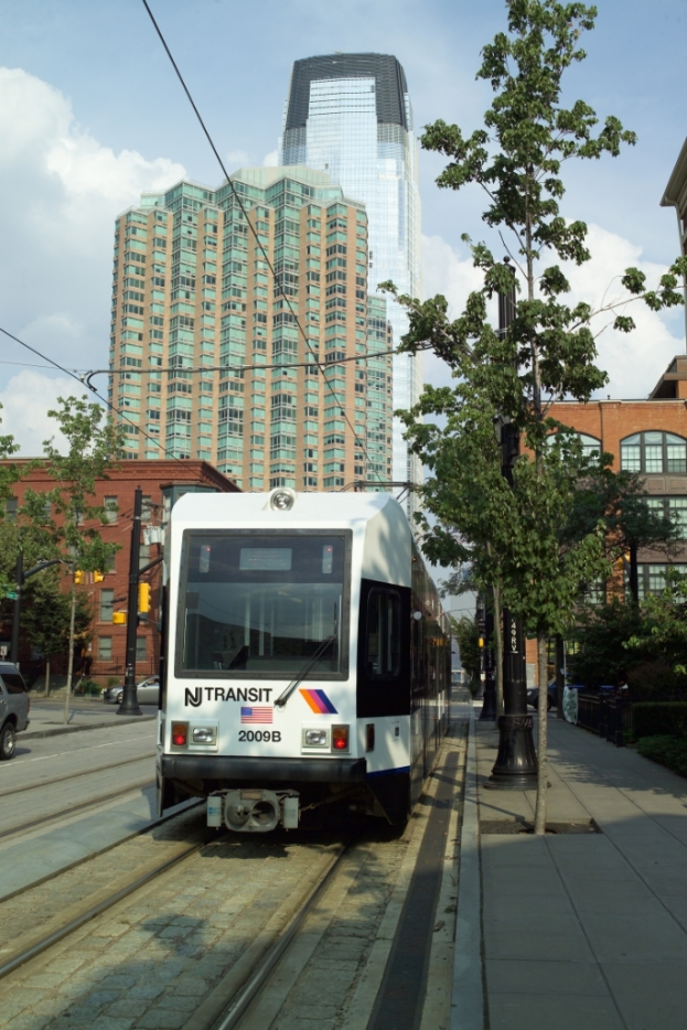 Ein Zug auf Schienen mit Oberleitungen, flankiert von Bäumen, Masten und Gebäuden, mit einem Wolkenkratzer und Himmel im Hintergrund.
