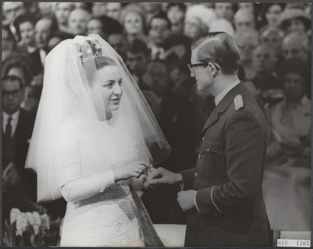 Ein Schwarz-Weiß-Foto eines Mannes und einer Frau, die sich die Eheringe reichen, mit einer Gruppe von Menschen im Hintergrund.