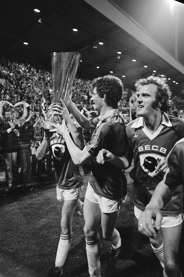 Schwarzes und weißes Foto von Fußballspielern, die einen Pokal auf einem Feld halten und feiern, während Zuschauer von hinter einem Zaun zusehen.