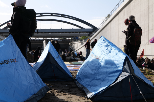 Eine Gruppe von Menschen sitzt auf einem sandigen Strand in der Nähe von Zelten, mit einer Wand und einer Brücke im Hintergrund, die Gegenstände während einer Klimawandel-Demonstration halten.