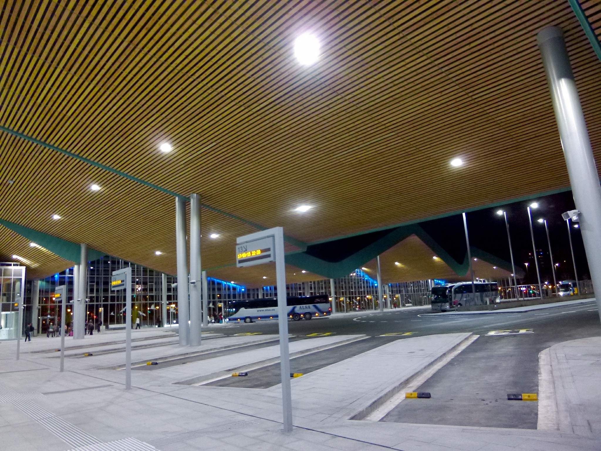 Bus-Haltestelle bei Nacht mit Deckenbeleuchtung, Fahrzeuge auf der Straße, Pfosten, Texttafeln, einige Menschen und beleuchtete Gebäude im Hintergrund.