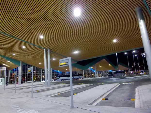 Bus-Haltestelle bei Nacht mit Deckenbeleuchtung, Fahrzeuge auf der Straße, Pfosten, Texttafeln, einige Menschen und beleuchtete Gebäude im Hintergrund.