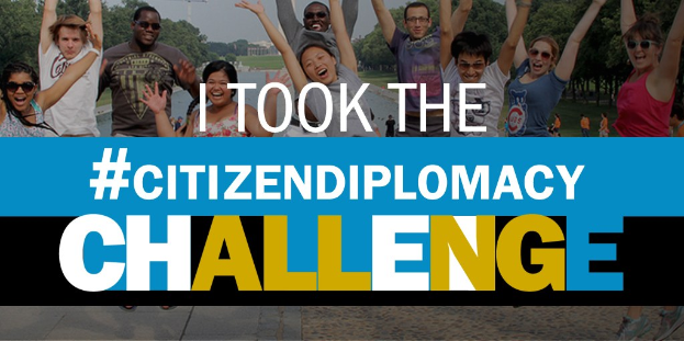 Eine Gruppe von Menschen, die vor einem Schild mit der Aufschrift "Ich habe die Staatsbürgerschaftsherausforderung angenommen" lächeln und posieren, mit Bäumen, Gebäuden, Kränen und einem klaren blauen Himmel im Hintergrund.