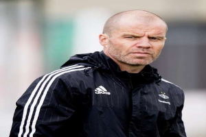 Kahlköpfiger Trainer in marineblauer Jacke mit weißen Streifen und weißem Kragen auf einem Fußballfeld stehend, die Hände verschränkt und einen ernsten Gesichtsausdruck tragend.