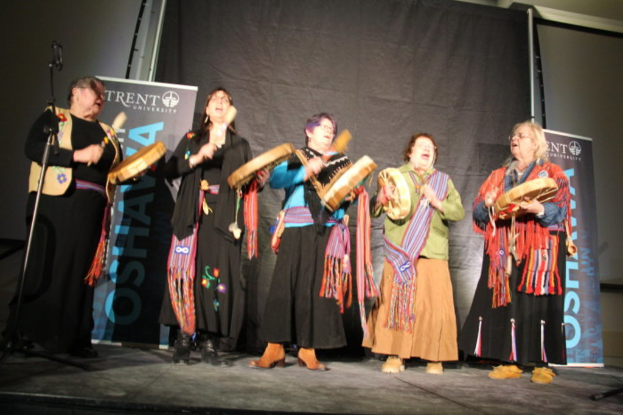 Eine Gruppe von Frauen in bunten Kleidern, die singen und Trommeln halten, vor einem schwarzen Vorhang und zwei blauen Rollplakaten.
