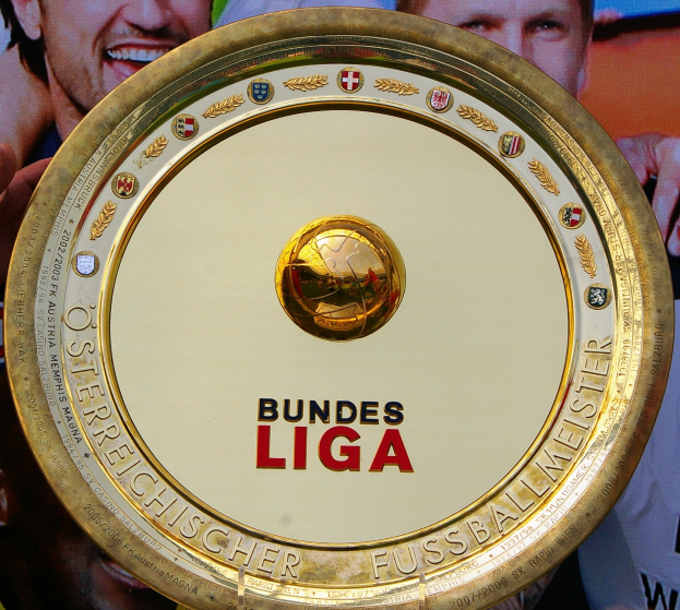 Ein Mann hält einen goldenen Schild mit der Aufschrift 'Bundesliga' und im Hintergrund eine Fahne mit drei Personen.