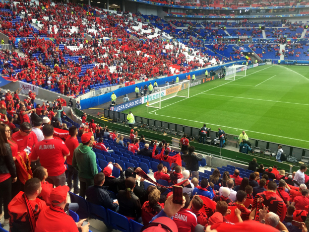 Eine große Menge Menschen in einem Stadion schaut einem Fussballspiel zu, mit Menschen auf Sitzen und Stehplätzen, Bannern, einem Torpfosten und Deckenbeleuchtung.