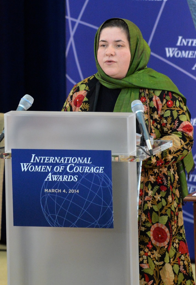 Eine Frau steht an einem Rednerpult mit einem Mikrofon, einer Tafel und einem Tisch rechts daneben, mit einem Banner im Hintergrund mit der Aufschrift "International Women of Courage Awards 2014".
