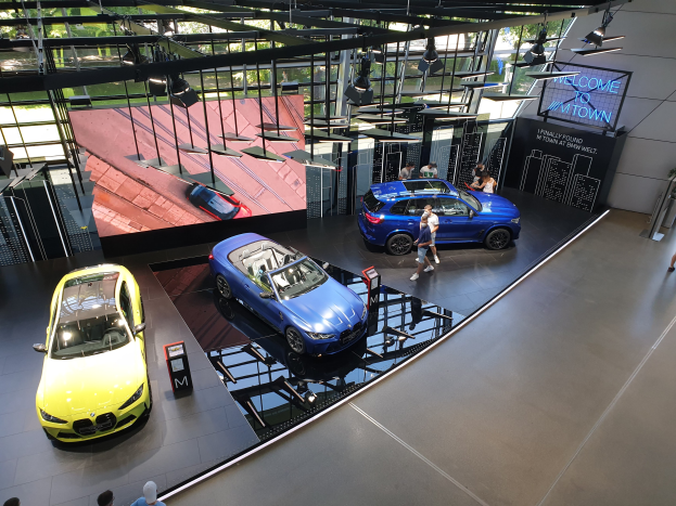Gruppe von Menschen auf dem Boden des BMW Museums in München, Deutschland, mit Autos zur Linken und einem "BMW Museum München"-Schild zur Rechten, mit Blick auf Bäume, Gebäude und den Himmel durch Glasfenster.