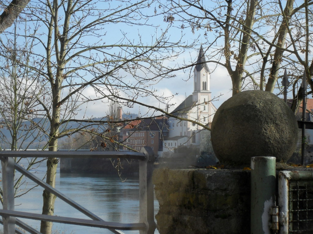 Ein hoher Pfeiler steht vor Gebäuden mit Fenstern, Bäumen mit Ästen, einem Tor und fließendem Wasser.
