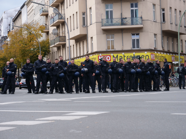 Eine Gruppe von Polizisten in schwarzen Uniformen hält blaue Helme in der Mitte einer Straße, die von Laternenmasten, Bäumen und Gebäuden mit Glasfenstern und Balkonen gesäumt ist, mit mehr Bäumen und einem klaren blauen Himmel im Hintergrund.