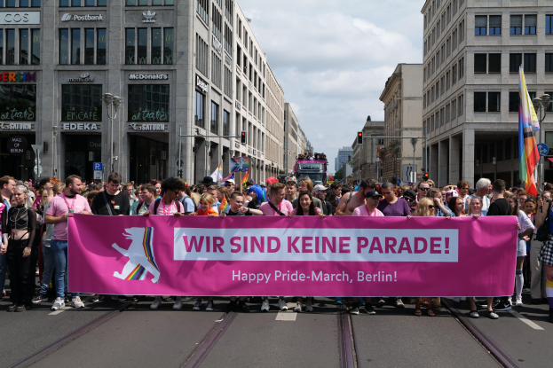 Eine Gruppe von Menschen marschiert auf einer Straße in Berlin, Deutschland, mit einer pinken "Happy Pride March"-Fahne, während Gebäude, Laternenpfähle und Verkehrszeichen die Straße säumen, unter einem bewölkten Himmel.