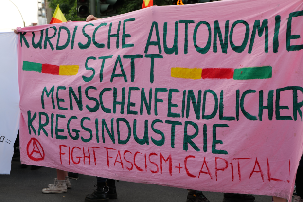 Eine Gruppe von Menschen steht auf einer Straße und hölt ein pinkes Banner mit der Aufschrift "Kurdische Autonomie Statt Menschenfeindlicher Kriegsindustrie Fight Fascism Capital", mit Ampeln, Flaggen, Bäumen, Gebäuden und einem klaren blauen Himmel im Hintergrund.