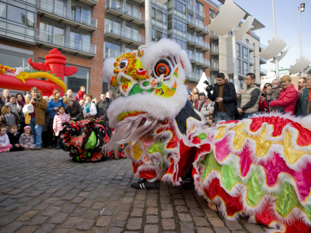 Lebendige Feier des chinesischen Neujahrs in Amsterdam mit einer Löwen-Tanz-Performance vor einer Zuschauermenge, die einige Kameras halten, vor einer Kulisse aus Gebäuden, Laternenmasten und einem klaren blauen Himmel.