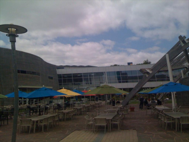 Eine Resortszene mit Stühlen, Tischen, Sonnenschirmen und einer Lampe im Vordergrund, Bäume und Gebäude in der Mitte, unter einem bewölkten Himmel.