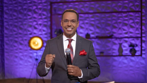 Stephen Colbert, Moderator von "The Daily Show with Trevor Noah", steht auf einer Bühne und hält ein Mikrofon in der Hand, trägt einen Anzug, eine Krawatte und eine Brille, mit einer Wandmontage und einem Licht hinter ihm.