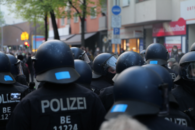 Eine Gruppe von Polizisten in schwarzen Uniformen und Helmen steht vor einer unscharfen Menge und einer Stadtlandschaft mit Gebäuden, Bäumen, Pfählen, Verkehrszeichen und Schildern.