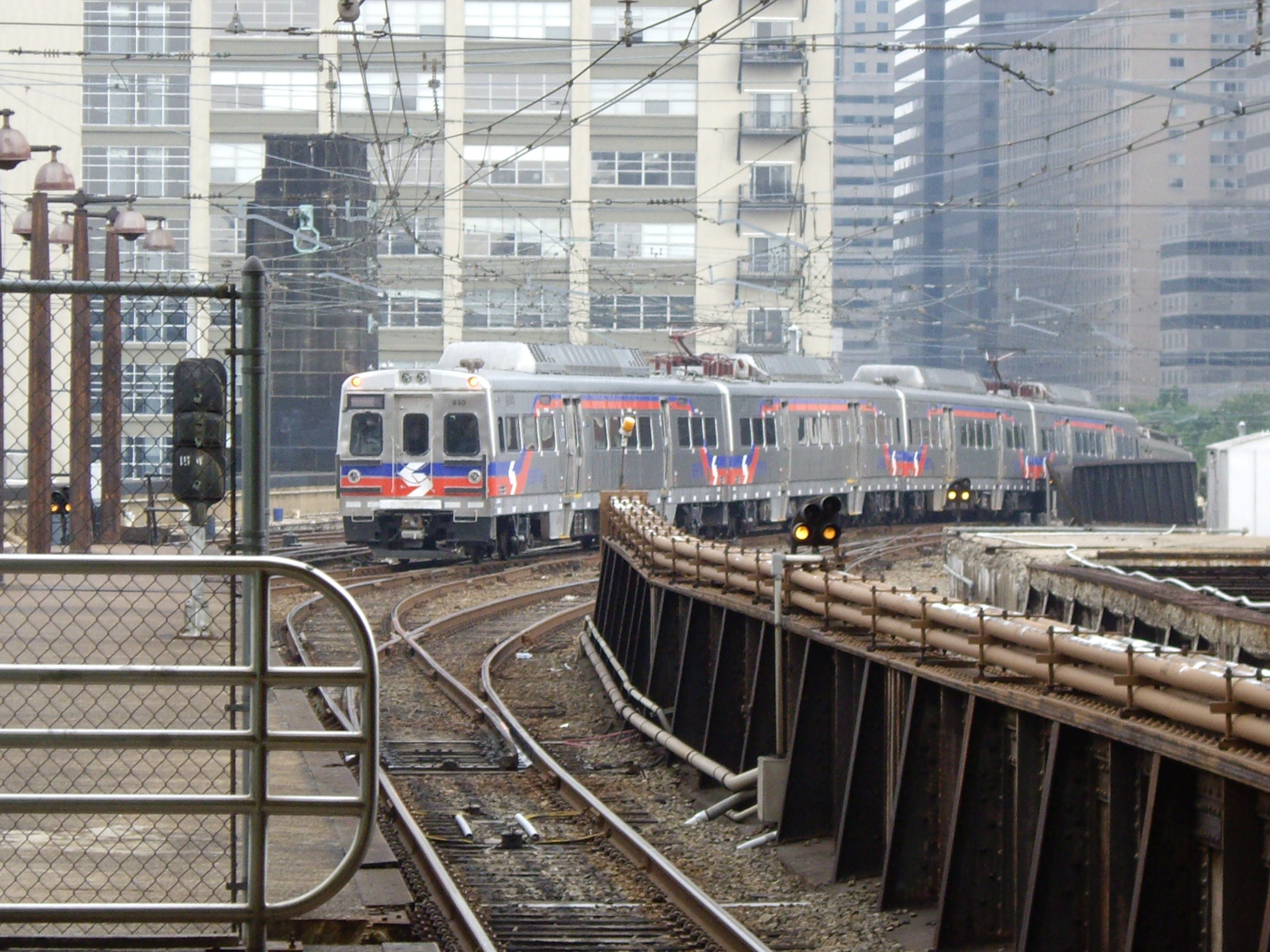 Nahverkehrszug fährt auf Gleisen neben Hochhäusern mit städtischer Infrastruktur und Grünflächen im Hintergrund.